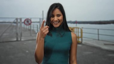 Okyanus manzaralı deniz kıyısında gülümseyen ve parmaklarına güvenen bir kadın bulutlu bir gökyüzünün altında neşeli bir ruh hali olduğunu gösterir..