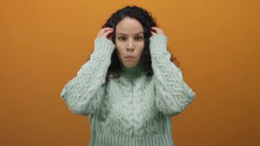 Yeşil kazaklı, kıvırcık saçlı genç bir kadın turuncu bir arka plan karşısında şaşkınlığını dile getiriyor..