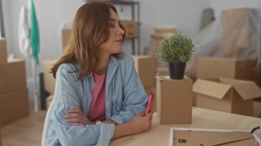 Genç bir kadın yeni bir evde oturuyor. Etrafı kutularla çevrili, elinde telefon, ve aydınlık bir oturma odasında mutlu görünüyor..