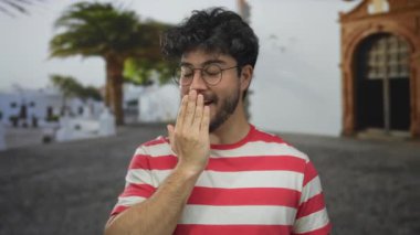 Çizgili tişörtlü, sakallı genç adam güneşli bir sokakta ağzı şakacı bir şekilde kapatıyor. Avuç içi ve kemer arkası da sürpriz ya da gizliliği gösteriyor..