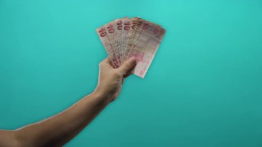 Ekonomi ve finans kavramını vurgulayarak, izole edilmiş mavi bir zemin karşısında Türk lirası banknotları tutan bir adamın eli.