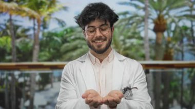 Sakallı ve gözlüklü genç adam güneşli bir parkta gülümsüyor beyaz bir ceket giyiyor, yemyeşil palmiye ağaçlarıyla çevrili, açık elleri ve sakin tavırları var..