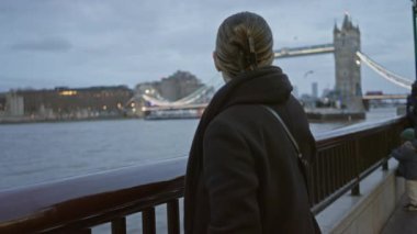 Londra 'da gülümseyen kadın nehir kıyısında duruyor. Arka planda köprü var. Bir akşam manzarası yakalıyor..