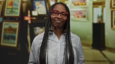 Gözlüklü ve uzun örgülü bir kadın sanat galerisinde jest yapıyor, çerçeveli sanat eserleriyle çevrili, kendinden emin ve dışa dönük tavırlar sergiliyor.