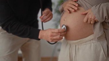 Hamile kadın ve erkek odada, steteskopla bebeğin kalp atışlarını dinliyorlar, ev ortamında samimi bir an yaratıyorlar, hamileliğin altını çiziyorlar ve çiftlerin arasında bir bağ kuruyorlar..