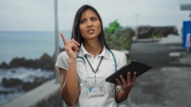 Steteskoplu ve tabletli kadın doktor deniz kenarında sakin bir arka planda gezinirken açık hava ortamında tıbbi danışmanlığı temsil ediyor.