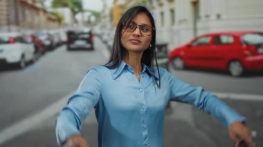 Mavi gömlekli Latin kadın, kırmızı arabalarla ve arka planda bulanık binalarla, gençlik enerjisi ve kentsel ortamları somutlaştırarak, şehir caddesinde heyecana işaret ediyor..