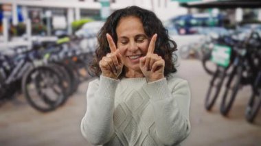 Kıvırcık saçlı, gülümseyen ve kalp şeklinde parmaklarını açık havada bisiklet kiralama istasyonunun yanında gezdiren park halindeki bisikletlerle ve canlı şehir manzarasıyla çevrili bir kadın..