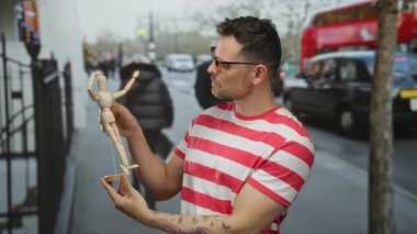 Gözlüklü ve çizgili tişörtlü genç adam şehir caddesinde ahşap bir manken tutuyor. Arka planı bulanık insanlar ve arabalar yolda ilerliyor..