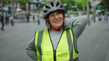 Güvenlik yeleği ve miğferi olan yaşlı İspanyol kadın şehir sokaklarında dikiliyor ve gergin şehir ortamındaki gerginlik ve gerginlikten dolayı boynunu ovalıyor..