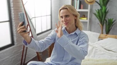 Pijamalı bir kadın parlak yatak odasında akıllı telefondan sohbet ediyor ve pencereden ışık süzülüyor..