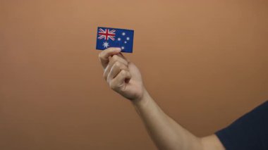 Kahverengi duvara karşı küçük Avustralya bayrağı taşıyan bir adam nakış işine odaklanmış, ulusal gururunu ve kimliğini basit ve izole bir ortamda sergiliyor..