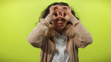 Kıvırcık saçlı, dürbünü canlı sarı arka planda taklit eden kadın neşeli bir merak ve neşe yayar..