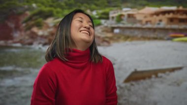 Açık havada gülümseyen, deniz manzaralı kırmızı süveter giyen bir kadın doğal bir kıyı ortamında yemyeşil sakin bir sahil manzarası sergiliyor..