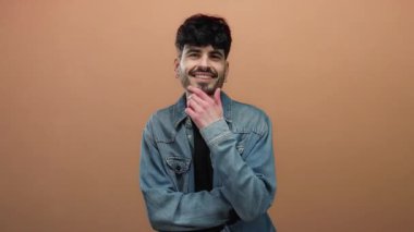 Kot ceketli genç adam, düz kahverengi bir arka plan önünde kendinden emin bir şekilde gülümsüyor..