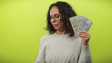 Gözlüklü orta yaşlı bir kadın, Amerikan doları banknotlarını canlı sarı bir arka plana karşı tutuyor, zenginlik ve güveni resmediyor..