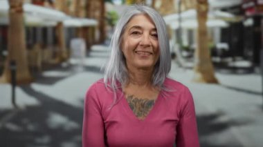 Gri saçlı yaşlı bir kadın güneşli bir sokakta taştan bir jest yapıyor pembe bir kazak giyiyor ve kendine güvenen ve mutlu bir ifadeyle gülümsüyor..