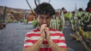 Sakallı genç adam açık hava kaktüs bahçesi parkında dua ediyor. Çizgili gömlek ve gözlük takıyor. Açık mavi gökyüzünün altında odaklanmış ve sakin görünüyor..