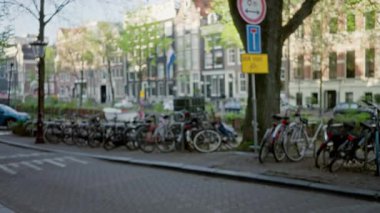 Amsterdam Canal Caddesi 'nin bulanık manzarası. Park edilmiş bisikletler, klasik Hollanda mimarisi ve parlak gökyüzünün altındaki yeşil ağaçlar..
