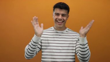 Genç adam elleriyle yüzünü kapatıyor, sonra turuncu bir arka plana gülümsüyor çizgili bir gömlek giyiyor, basit ama büyüleyici bir portrede neşe ve neşe ifade ediyor..