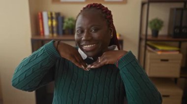 Kitaplık ve çiçeklerle dolu bir ofiste gülümseyen, yeşil bir kazak giyen, kapalı bir iş yerinde dostça bir yüz ifadesi sergileyen bir kadın..