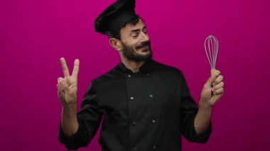 Siyah üniformalı İspanyol sakallı şef canlı pembe bir arka planda çırpı çırpıyor, iki eliyle barışı işaret ediyor, neşeli yemek pişirme ruhunu somutlaştırıyor..
