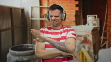 İnşaat alanında çizgili tişörtlü kulaklık takan, el hareketleriyle duygularını ifade eden, kolunda dövmeler olan bir adam..