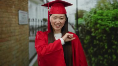 Kırmızı mezuniyet elbisesi giymiş bir kadın, arka planda yeşillik olan şehir sokağında kendine güvenerek gülümsüyor..