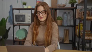 Ofiste dizüstü bilgisayarda çalışan ve gözlüklü kızıl saçlı bir kadın. Etrafı raflar ve bitkilerle çevrili, rahat bir profesyonel ortam yaratıyor..