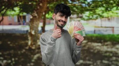 Parkta elinde Norveç banknotlarıyla gülümseyen bir adam çevrenin yeşillikle çevrili olduğunu gösteren olumlu bir hareket sergiliyor..