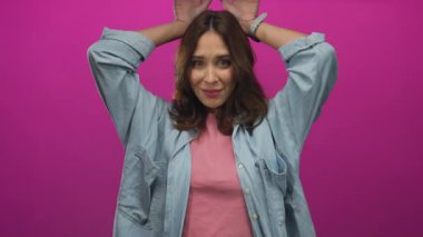 Genç İspanyol kadın neşeli bir şekilde tavşan kulağı hareketi yapıyor. Ellerini başının üstüne koyup, izole edilmiş pembe bir arka plana yaslıyor. Günlük kot ve pembe bluz giyiyor..
