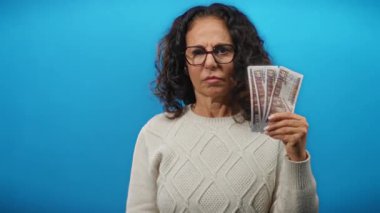 Orta yaşlı bir kadın elinde Mısır banknotları, kulağını mavi duvara dayamış, hayal kırıklığı ve finansal gerginlik gösteriyor..