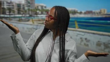 Gözlüklü kadın plaj gezintisinde huzur içinde poz veriyor. Arka planda deniz kenarı var. Kıvırcık saçlar, sakin bir ifade ve sessiz tonlar sergiliyor..