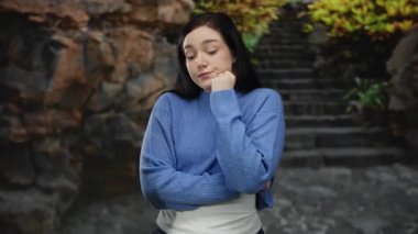 Kadın düşünceli bir şekilde bir parkta durmuş yeşil ışığın altında, taştan merdivenler ve yemyeşil bir yeşillik zeminine kurulmuş hayatı düşünerek, açık hava güzelliğini somutlaştırıyor..