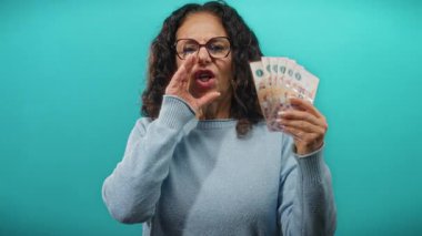 Arjantinli pezolarla gülümseyen ve bağıran bir kadın izole edilmiş mavi bir duvarın üzerinde bir anlık heyecan ve finansal refah yakalıyor..