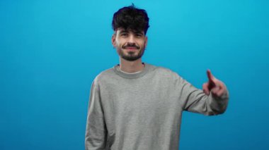 Genç ve yakışıklı bir adamın portresi gülümseyen ve izole edilmiş mavi bir arka plan üzerinde barış işareti yapan, kendine güveni ve arkadaşlığı şık bir kıyafetle yayan.