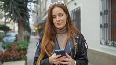 Kadın şehir caddesinde akıllı telefon kullanıyor, deri ceket giyiyor, şehir tarzını ve günlük modasını gün içinde sergiliyor..