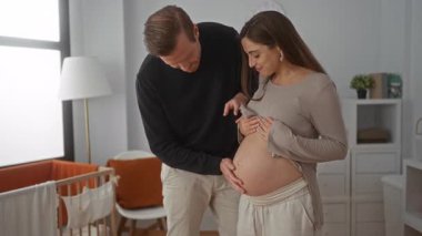 Bebek odasında hamile bir kadın ve erkek karnının üstünde hamileliği kucaklıyor, sıcak bir odada sevgi ve beklenti sergiliyor..