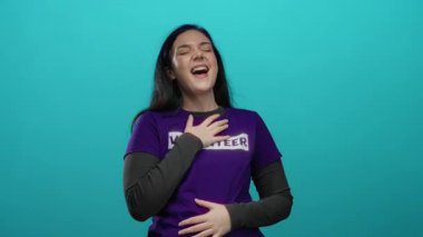 Mor gönüllü gömleğiyle mavi arka plana karşı tutkuyla şarkı söyleyen kadın, neşe ve duyguyu ifade ediyor, esmer, uzun saçlı ve elleri göğsünde.