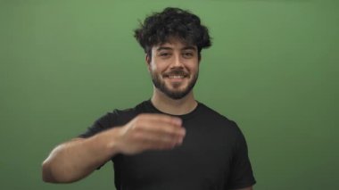 Sakallı genç İspanyol bir adam neşeli bir şekilde parmaklarıyla bir öpücük jesti gönderiyor. Canlı yeşil bir arka plan, neşeli bir ifade ve gündelik bir tarz sergiliyor..