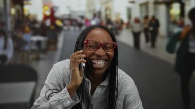 Akıllı telefonda konuşan bir kadın hareketli bir sokak terasında gülümsüyor canlı şehir hayatını ve sosyal etkileşimi gözler önüne seriyor..