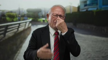 İş elbisesi giymiş kıdemli bir adam sokakta dikiliyor, burnunu eliyle kapatıyor, rahatsızlık tasvir ediyor, gündüz vakti şehir manzarası ile çevrili..