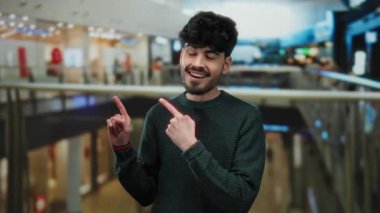 Genç adam, hareketli bir alışveriş merkezini işaret ederek canlı bir alışveriş havası ve şaşkınlık ve merak ifadeleri sergiliyor..
