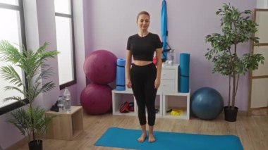 Spor salonunda yoga minderiyle egzersiz yapan bir kadın. Etrafı spor aletleri ve bitkilerle çevrili. Kapalı alanda rutin sağlık yaşam tarzını sergiliyor.