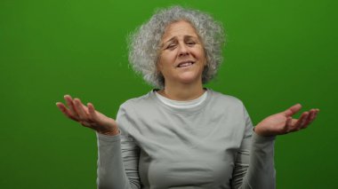 Gri saçlı bir kadın, yeşil arka plana karşı duygularını dışa vuruyor rahat ve derin derin bir duruşla, kapalı bir yerde gri bir bluz giyiyor..