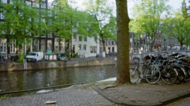 Ağaçların, motosikletlerin ve amfi kanalların bokeh görüntüsünü gösteren bulanık bir sahne, yemyeşil yaprakların altındaki Hollanda suyolları boyunca uzanan açık hava sükunetini vurguluyor.