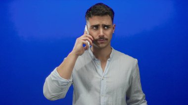 Genç İspanyol asıllı bir adam, mavi arka planda akıllı telefondan konuşuyor, çılgın jestler yapıyor, rahat bir gömlek giyiyor, hayal kırıklığını ifade ediyor, yüz ifadeleriyle duygularını iletiyor..