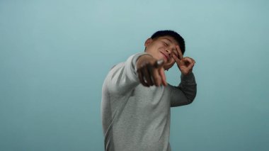 Genç adam güvenle gülümsüyor ve özgüvenli bir mavi arka plana işaret ediyor, bir stüdyo ortamında neşeli ve çekici bir tavır yakalıyor..