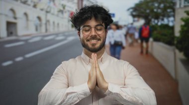 Gözlük takan sakallı genç adam, kalabalık bir sokakta dua jestinde dışarıda dururken gülümsüyor.