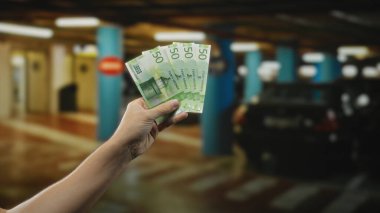 Elinde Norveç kronu banknotları tutan adam loş bir park alanında bulanık arabalarla kapalı bir ortamda mali ya da ticari bir tema öneriyor..